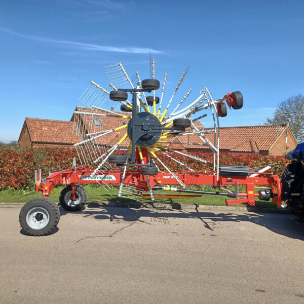 Red farm rake attached to a trailer being pulled by a blue tractor in a neighborhood yard under a blue sky. The machinery has many curved tines and black tires, with brick houses in the background.