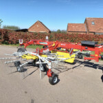Red agricultural implement with metal tines mounted on a trailer, parked in a driveway in front of a red-hedge and brick-tiled houses under a clear blue sky.