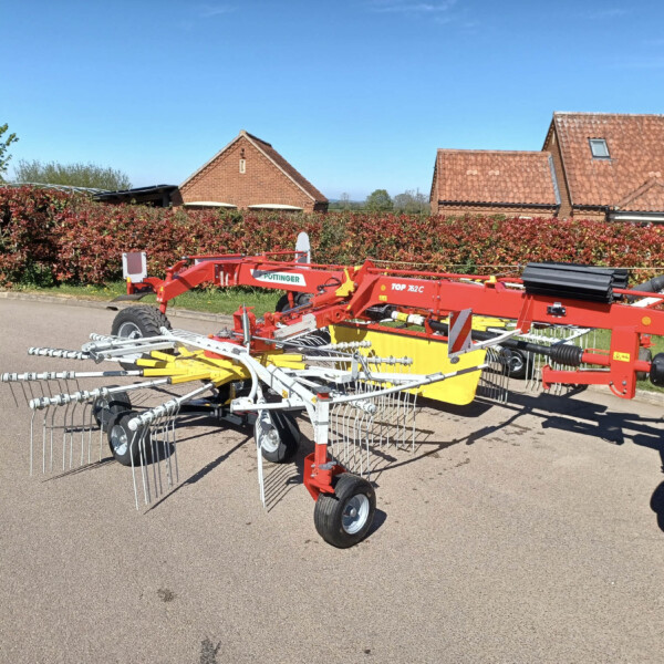 Red agricultural implement with metal tines mounted on a trailer, parked in a driveway in front of a red-hedge and brick-tiled houses under a clear blue sky.