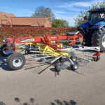 Blue tractor with large rear tire attached to red farming implement parked on a driveway beside a red-brick house and red hedge under a blue sky