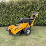 Yellow compact road roller parked on a grassy area with a dense green hedge in the background, outdoors.