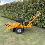 Yellow hedge-trimming machine parked beside a dense green hedge along a grassy area.
