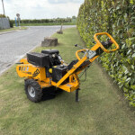 Yellow compact skid-steer loader parked on a grassy verge beside a hedge along a road, with a black tool basket on the front.