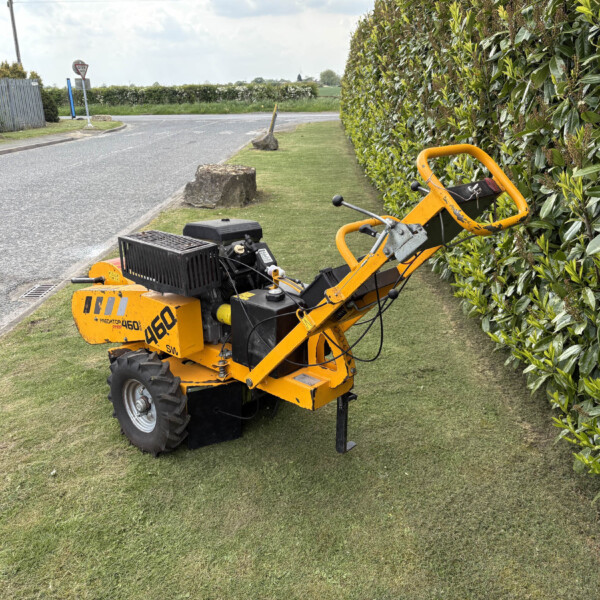 Yellow compact skid-steer loader parked on a grassy verge beside a hedge along a road, with a black tool basket on the front.