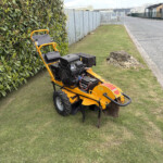 Yellow ride-on lawn mower parked on a grassy verge beside a dense hedge and a metal fence along a residential street, with houses in the distance.