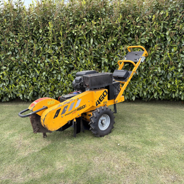 Small yellow ride-on construction machine parked on grass with a dense green hedge in the background.