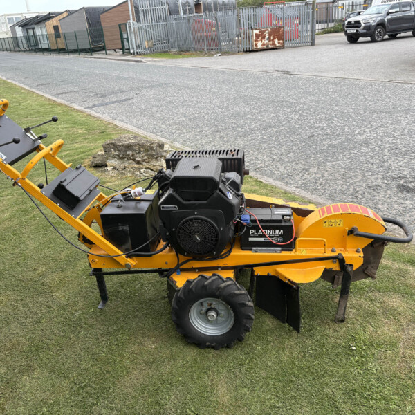 Yellow industrial riding mower parked on a grassy verge beside a paved road, with a chain-link fence and row of storage units in the background