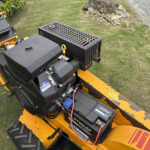 Close-up of a yellow lawn machine's engine area with a black battery box and exposed wiring on grass outside