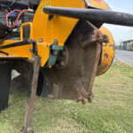 Close-up of a yellow road roller's front drum and rusted metal components, with wiring and black panels visible nearby on grass.