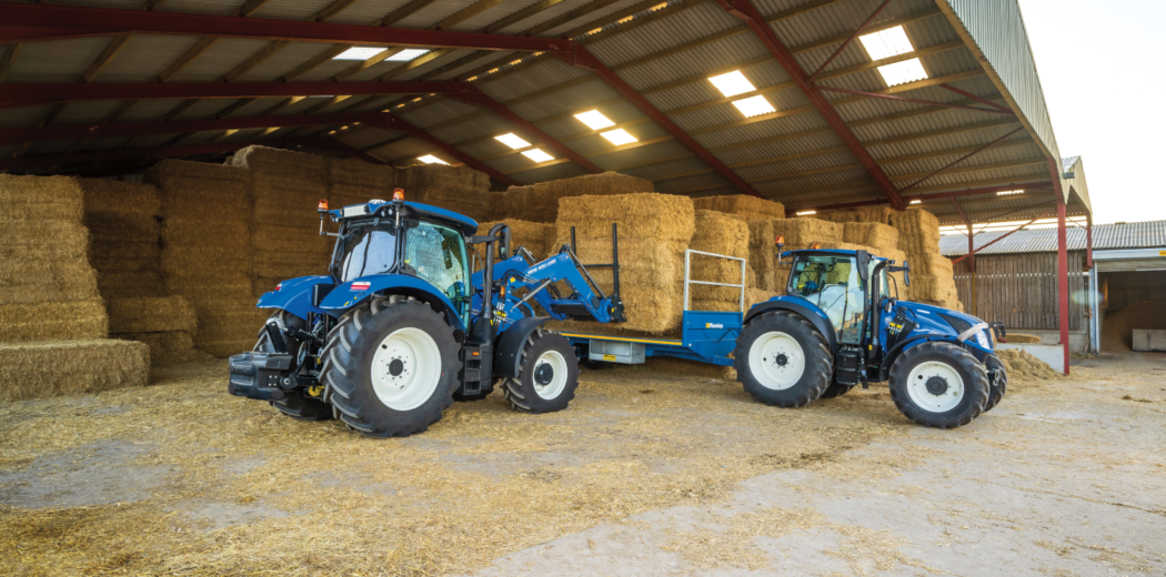 Two blue tractors with a hay bale trailer inside a barn, stacked hay bales in the background and dirt floor.