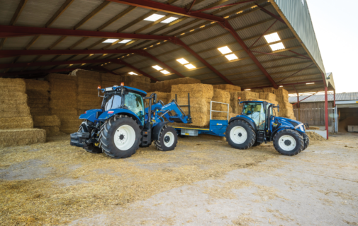 Two blue tractors with a hay bale trailer inside a barn, stacked hay bales in the background and dirt floor.
