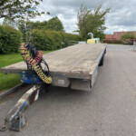 Flatbed trailer with coiled red, yellow, and black hoses attached, in a yard with hedges and trees and construction equipment in the background.