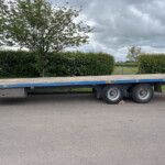 Blue flatbed trailer parked beside a hedge with trees and a cloudy sky in the background.
