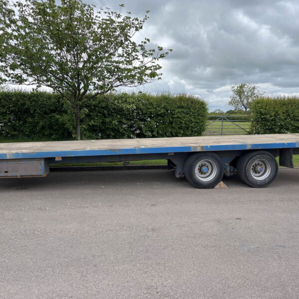 Blue flatbed trailer parked beside a hedge with trees and a cloudy sky in the background.