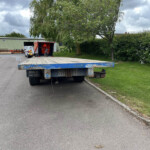 Blue flatbed trailer on an asphalt road in an industrial yard, with a green warehouse, a white van, red equipment, and trees in the background under cloudy skies, (image shows parked equipment).