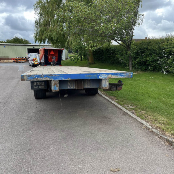 Blue flatbed trailer on an asphalt road in an industrial yard, with a green warehouse, a white van, red equipment, and trees in the background under cloudy skies, (image shows parked equipment).