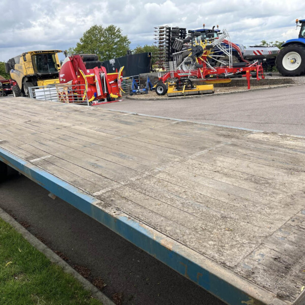 Flatbed trailer loaded with red agricultural equipment in a yard, with a blue New Holland tractor in the background under a cloudy sky.