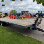 Farm equipment loaded on a flatbed trailer: yellow harvester, red baler, blue New Holland tractor, and hoses in the foreground under a cloudy sky.