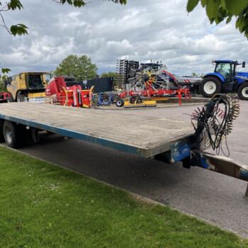 Farm equipment loaded on a flatbed trailer: yellow harvester, red baler, blue New Holland tractor, and hoses in the foreground under a cloudy sky.