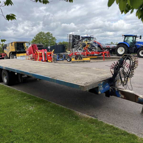 Farm equipment loaded on a flatbed trailer: yellow harvester, red baler, blue New Holland tractor, and hoses in the foreground under a cloudy sky.