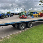 Row of blue tractors and green agricultural machinery on a flatbed trailer in a yard under a cloudy sky.