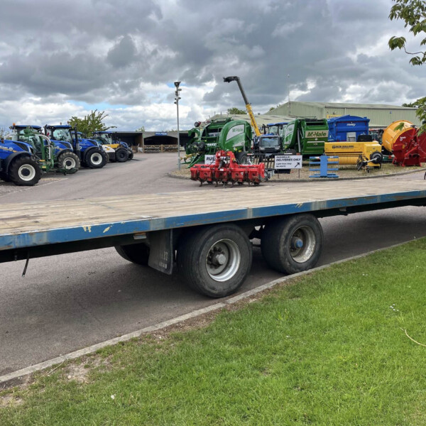 Row of blue tractors and green agricultural machinery on a flatbed trailer in a yard under a cloudy sky.