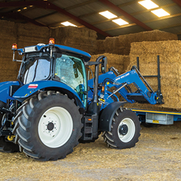 Blue tractor with front loader on a barn floor, loading stacked hay bales onto a trailer.