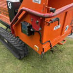 Close-up of an orange tracked construction machine showing hydraulic hoses and metal brackets on a grassy site area.