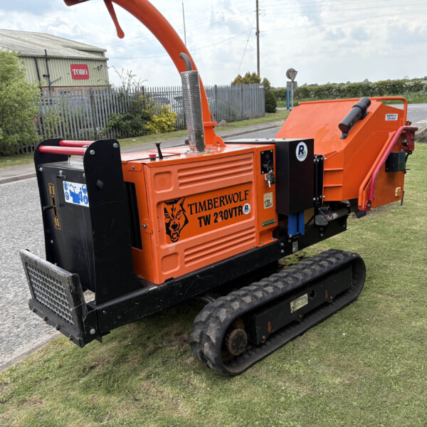 Large orange industrial wood chipper on tracked chassis beside a road, with metal chute and safety guards visible.