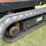 Close-up of a tracked vehicle's muddy metal track and undercarriage with an orange body, resting on grass.
