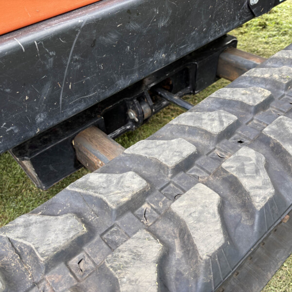 Close-up of a heavy machine's undercarriage with a muddy black track and orange body, resting on grassy ground.