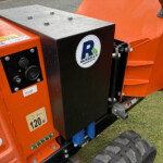 Orange industrial machine with a black control panel, featuring a Russell’s logo sticker and metal top plate with bolts, on a grassy surface.