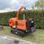 Orange Timberwolf trenching machine on tracks parked beside a hedge on a cloudy day.
