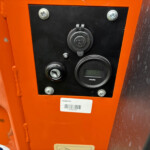 Close-up of an orange machinery control panel with a round 12V marine power outlet, screws, and an hour meter nearby
