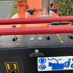 Close-up of orange construction vehicle deck with red guard rails and control panel stickers on a black surface.