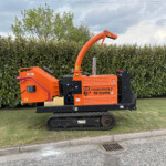 Orange Timberwolf TW 230VTR wood chipper on tracks beside a tall hedge, chute raised upward.