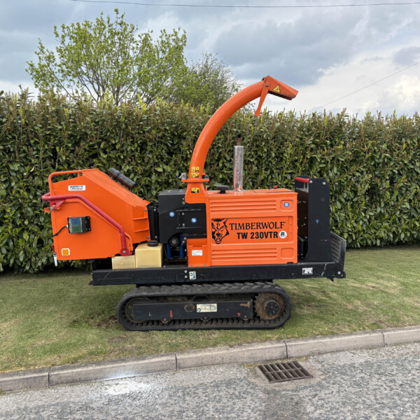 Orange Timberwolf TW 230VTR wood chipper on tracks beside a tall hedge, chute raised upward.