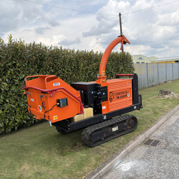 Orange Timberwolf TW 230TVR wood chipper on tracked undercarriage beside a hedge by a sidewalk, industrial buildings in the background.