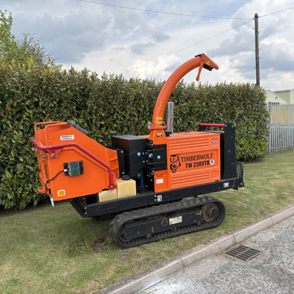 Orange Timberwolf wood chipper on tracks beside a hedge, with a metal fence and industrial building in the background.
