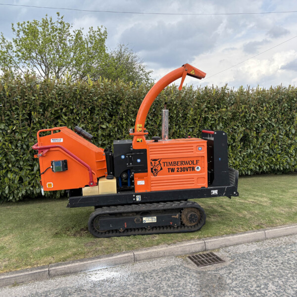 Orange Timberwolf TW 230VTR wood chipper on tracks, with a curved discharge chute, parked by a hedge on a grassy verge.