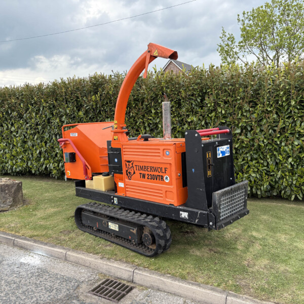Orange Timberwolf trenching machine on tracks parked beside a hedge on a cloudy day.