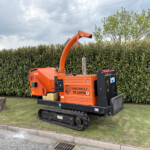 Orange Timberwolf wood chipper on tracks, parked beside a tall green hedge under a cloudy sky with its chute extended to the side.
