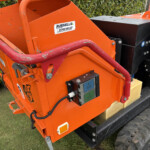 Close-up of an orange construction attachment with a red curved safety bar and a control module on a heavy vehicle.