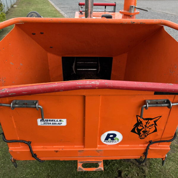 Orange construction skip from behind with chipped paint and stickers (Russells) visible; grassy verge on left and road on right, a person in dark clothing in the distance.