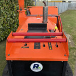 Orange industrial lift or crawler with control levers, orange rails, and perforated exhaust, parked beside a hedge and a sidewalk; Russell's branding on the front.
