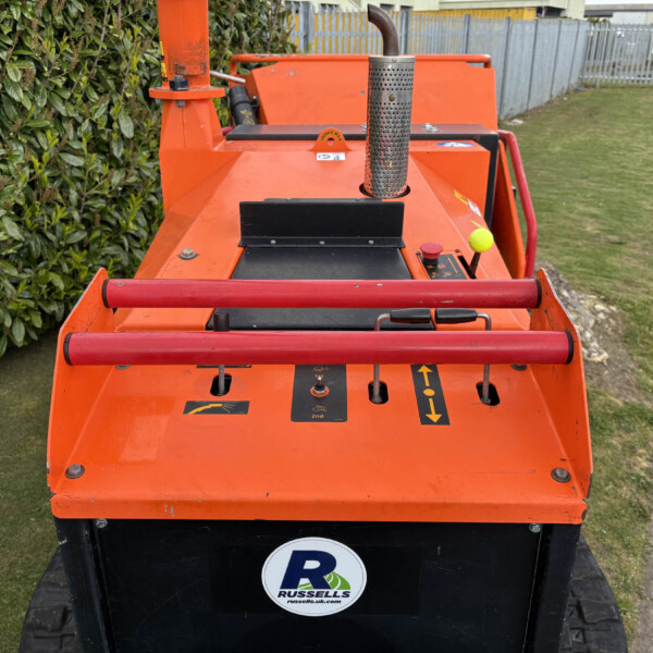 Orange industrial lift or crawler with control levers, orange rails, and perforated exhaust, parked beside a hedge and a sidewalk; Russell's branding on the front.