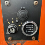 Close-up of an orange machine panel with a black control plate, showing a round 12V marine cap, rusted screws, and a gauge reading hours below the label Honeywell Quartz Hours.