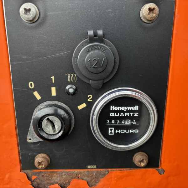 Close-up of an orange machine panel with a black control plate, showing a round 12V marine cap, rusted screws, and a gauge reading hours below the label Honeywell Quartz Hours.