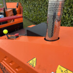Orange industrial machine with a perforated metal exhaust and warning stickers; a yellow tennis ball rests on the orange surface nearby.