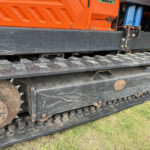 Close-up of a heavy construction vehicle's track system: orange body panel, black frame, and rusty metal tracks sitting on grass.